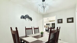 A dining area with a wooden table and chairs, a chandelier, and a kitchen in the background.