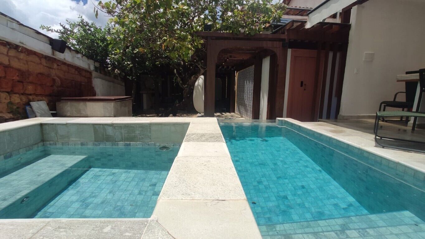 A rectangular swimming pool with clear blue water, surrounded by a stone wall and a wooden pergola.