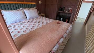A neatly made bed with floral bedding, a bedside table with a lamp, and a shelf with various items.