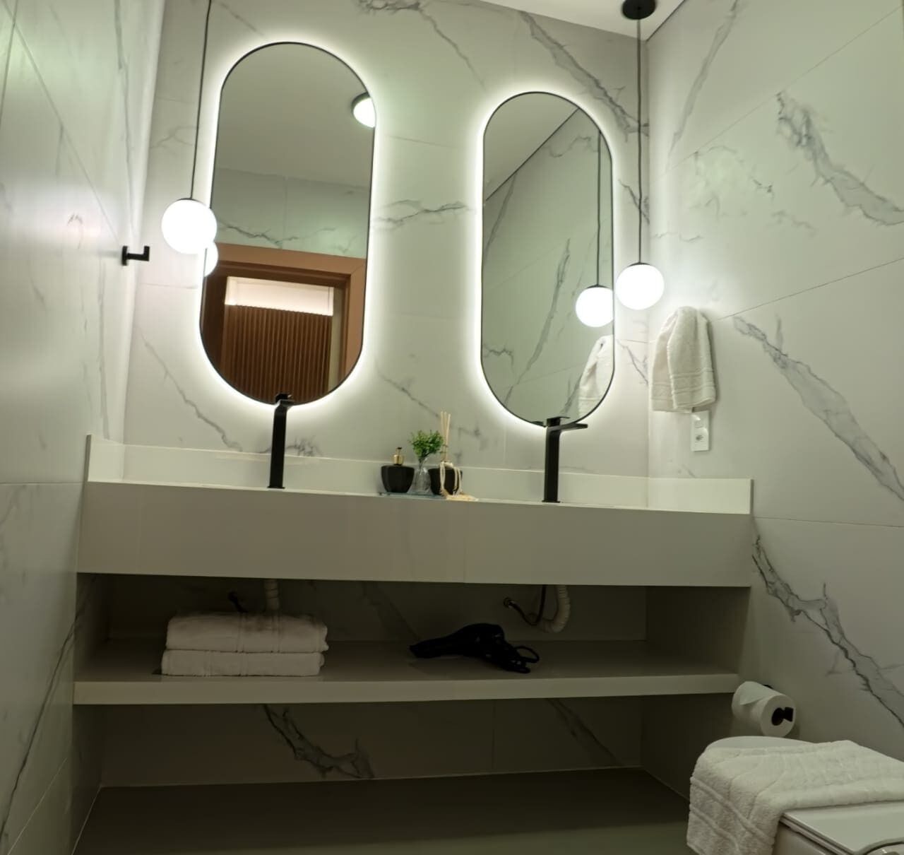A bathroom with a marble wall, a white vanity, two mirrors with lights, a towel rack, and a shelf with towels and a bottle.