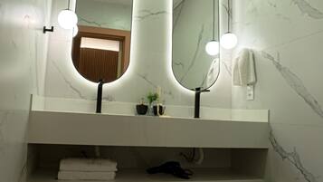 A bathroom with a marble wall, a white vanity, two mirrors with lights, a towel rack, and a shelf with towels and a bottle.