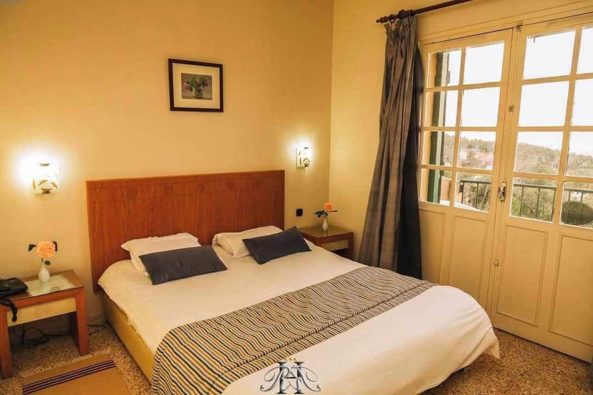 A hotel room with a large bed, two bedside tables, a nightstand, a framed picture on the wall, and a window with curtains.