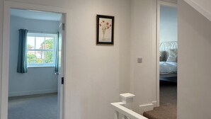 A staircase leading to a bedroom with a bed, a framed picture on the wall, and a door leading to a room with a window and curtains. - Modern 4-bed home near Thirsk| Ideal for Groups, Families & Work Stays (Rainton)