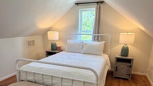 Un dormitorio con una cama, dos mesitas de noche, una ventana con cortinas, un radiador y un banco.