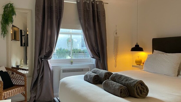 A hotel room with a bed, curtains, a bedside lamp, a plant, and a view of the outdoors. - Beautiful 2-bed Apartment In The Heart Of The City (Brighton and Hove)