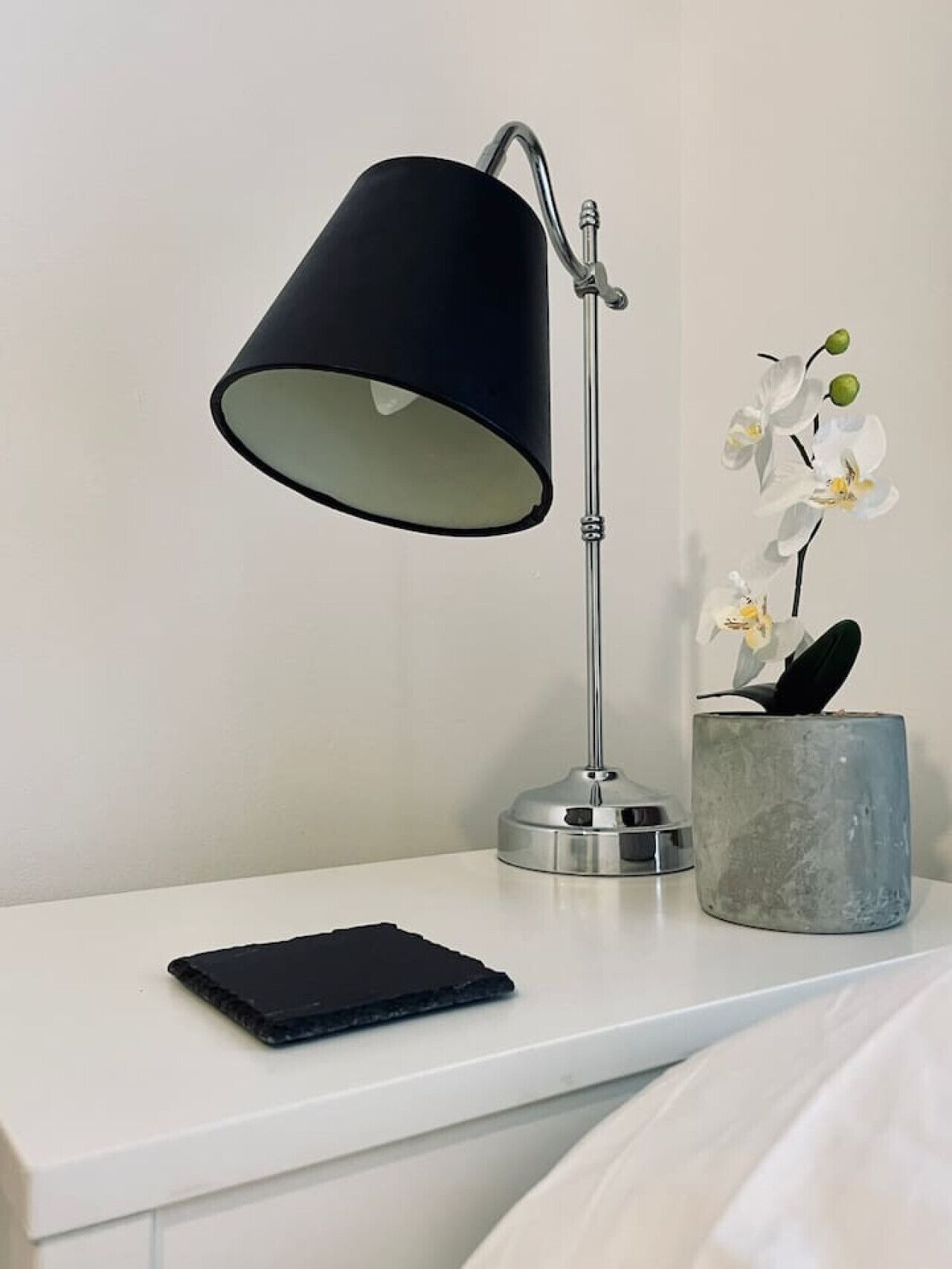 Une table de chevet avec une lampe de bureau noire, un téléphone intelligent et un vase contenant des fleurs blanches.