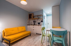 A compact living space with a yellow sofa, a small dining table with two chairs, a kitchenette with cabinets and a microwave, and a door leading to another room. - Welcome Flat in New Gudauri, Loft 1, RedCo (Gudauri)