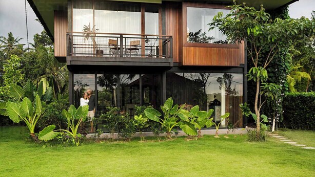 Une maison moderne à deux étages, dotée d’un balcon, entourée d’une végétation luxuriante et d’une pelouse bien entretenue.