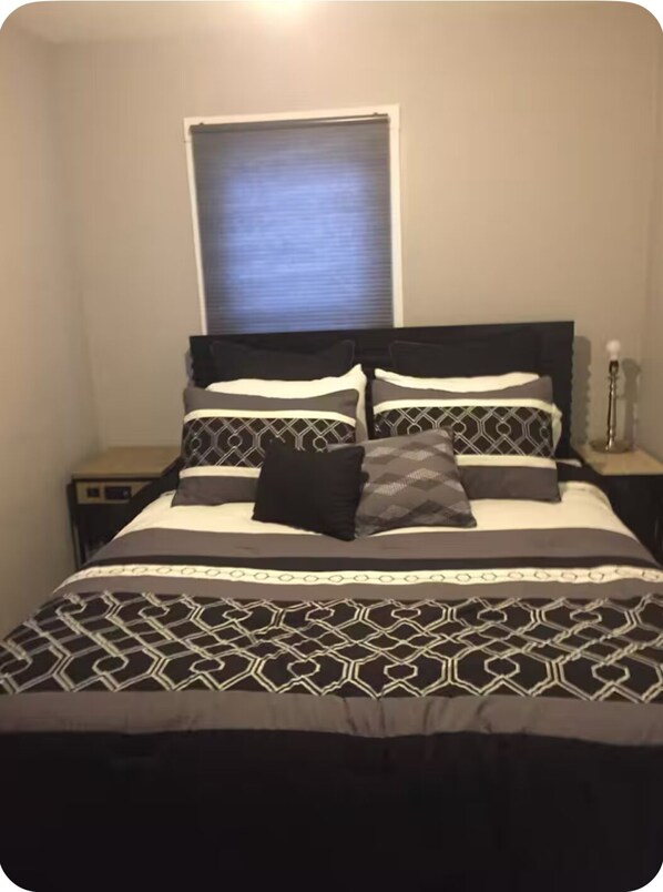 A bed with a patterned bedspread and pillows, a bedside table, and a window with blinds.
