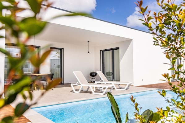 Un espace piscine extérieur moderne, doté de chaises longues, d’une table et d’un barbecue.