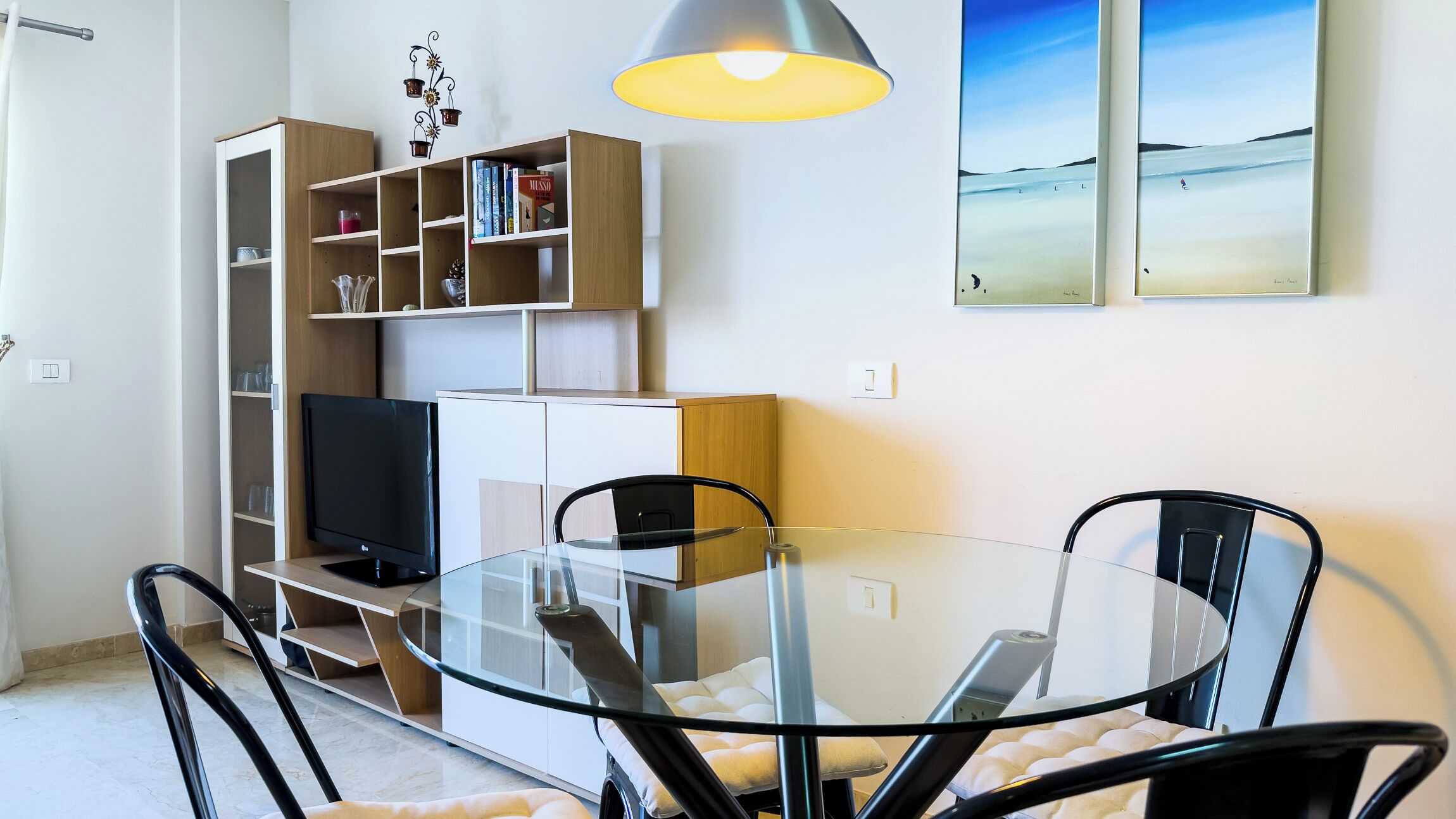 A modern living room with a glass dining table, black chairs, a TV, and a bookshelf.