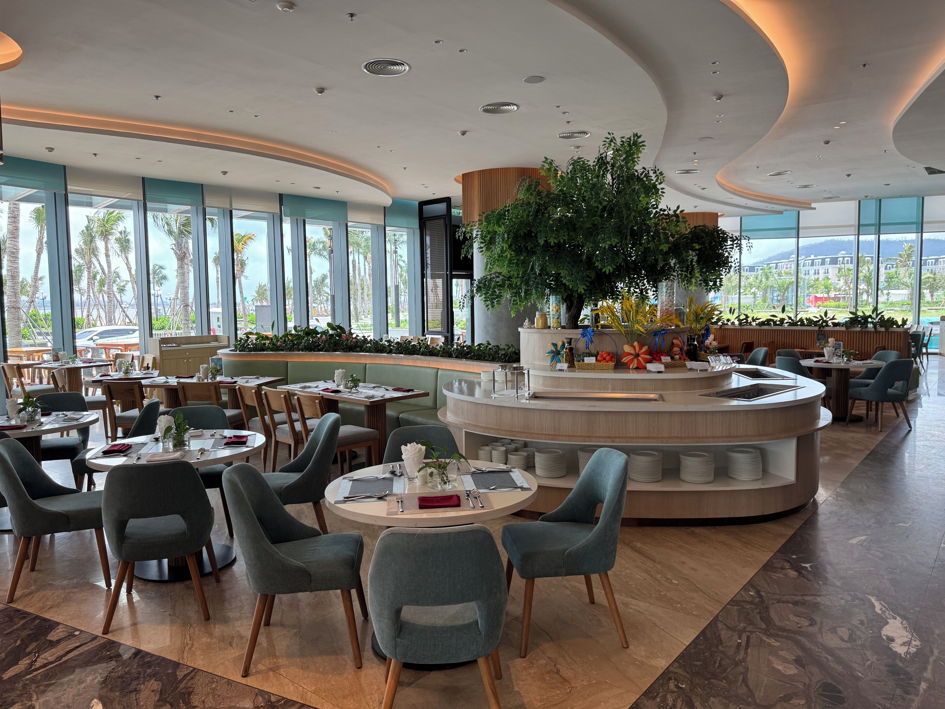 A modern restaurant with round tables, comfortable seating, a central decorative plant, and large windows offering a view of palm trees.