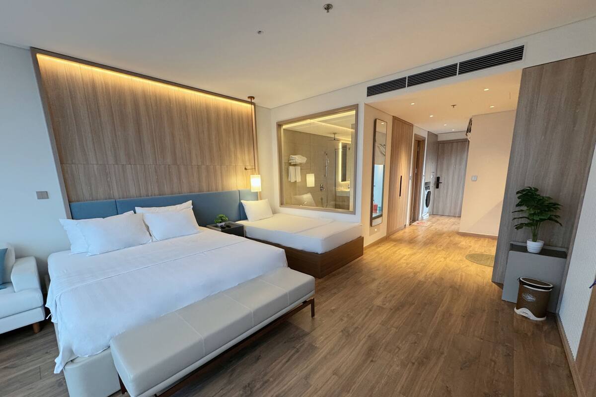 Quarto de hotel moderno com uma cama grande, uma cama menor, um espaço de estar e um banheiro visível através de uma porta aberta.