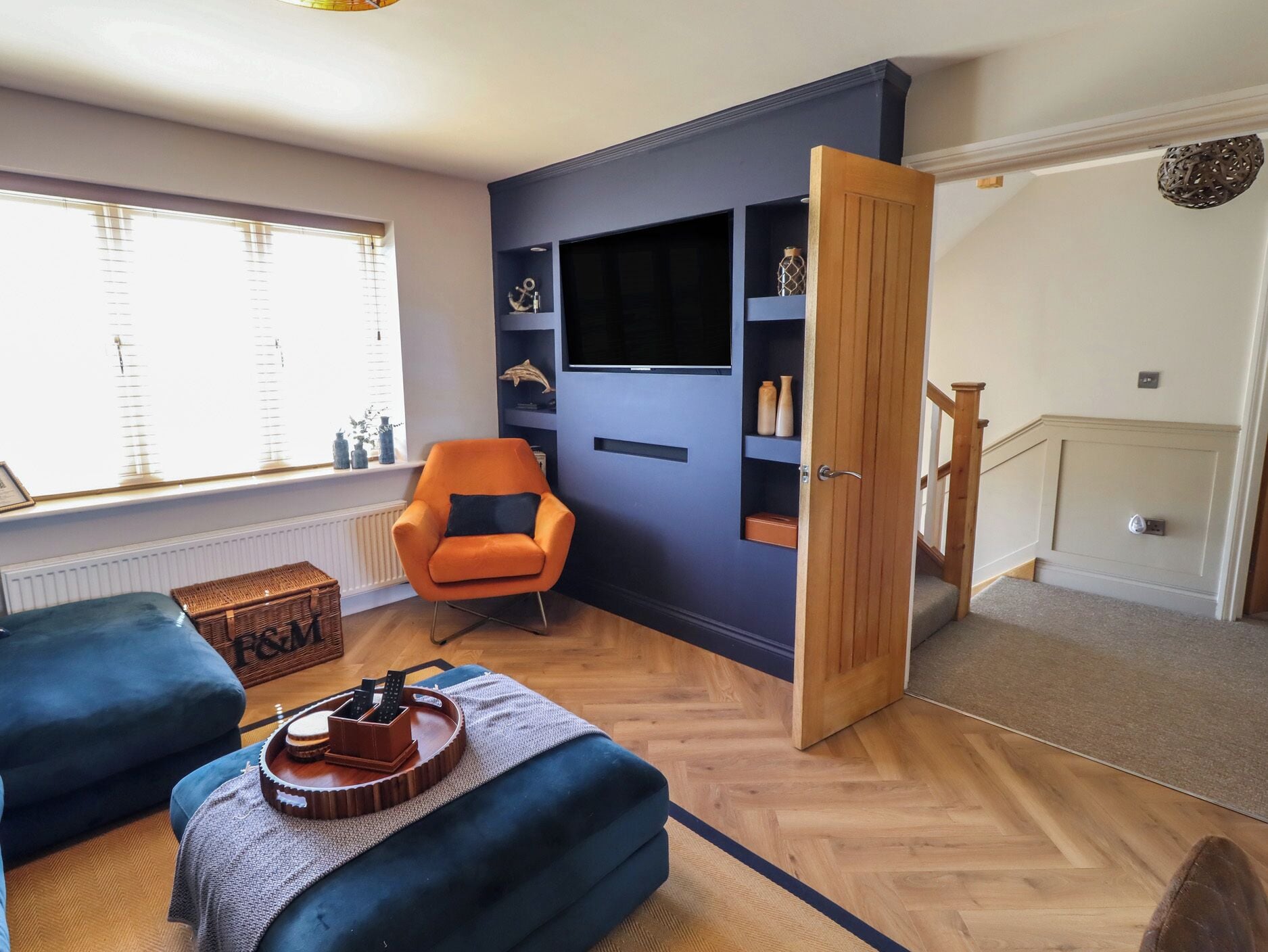 Une pièce avec un fauteuil bleu, une commode en bois, une petite table ornée d’un objet décoratif, un téléviseur accroché à un mur bleu et une porte en bois donnant sur une autre pièce.