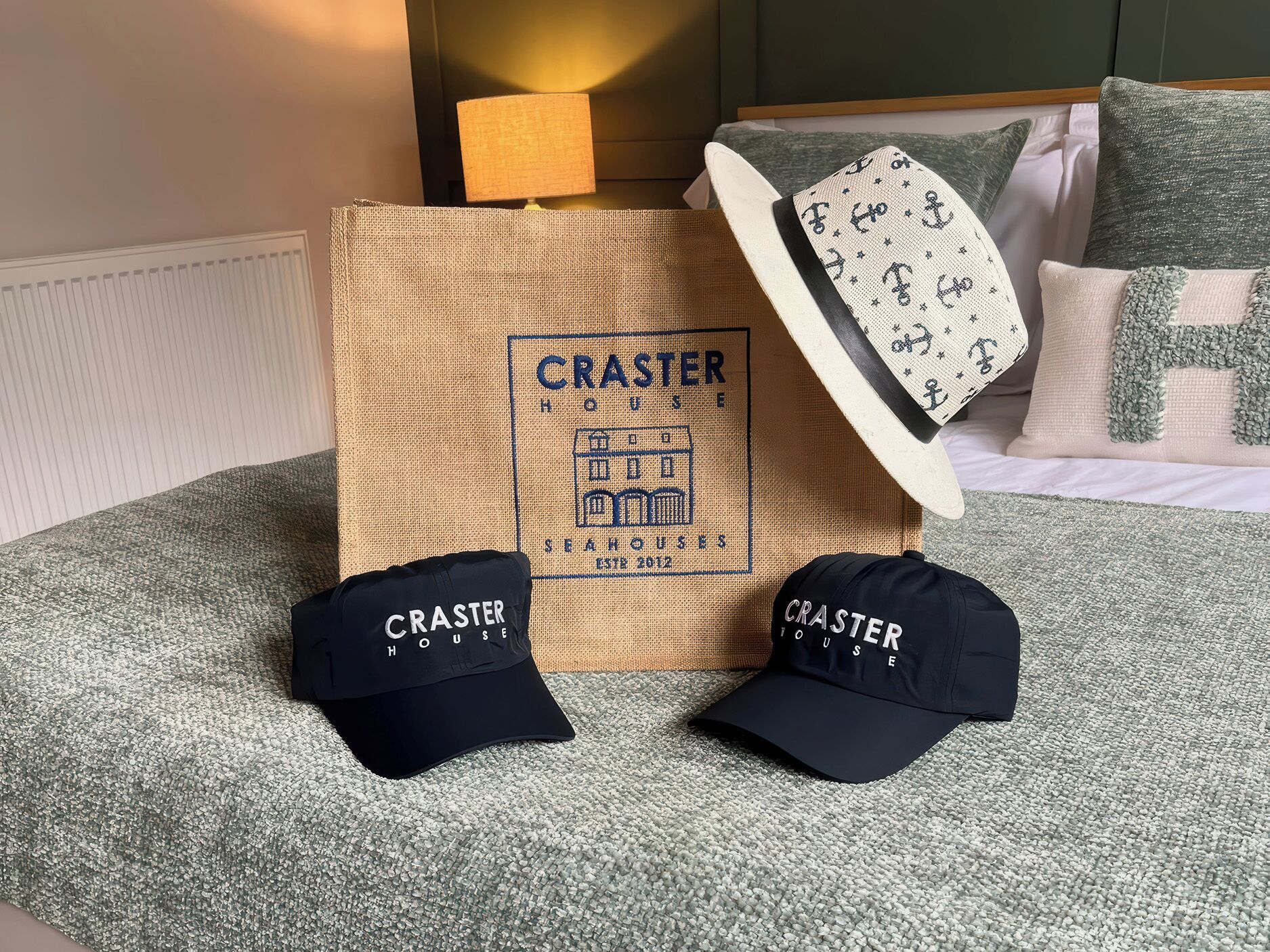 A bed with a green and white patterned pillow, a black and white cap, and a burlap bag with 'CRASTER HOUSE' branding.