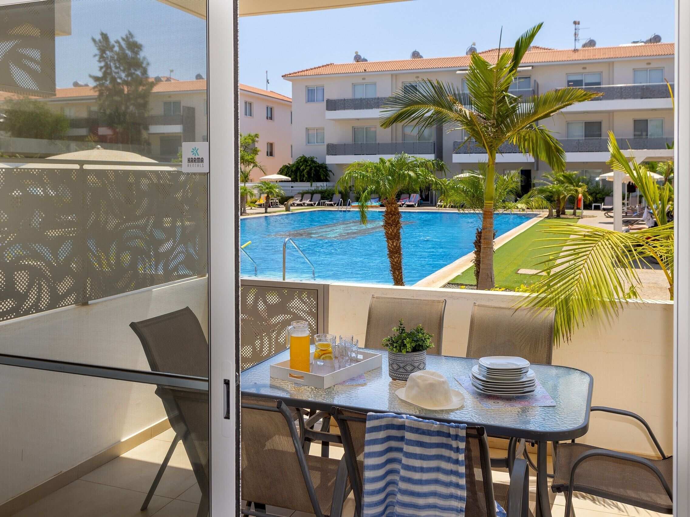 A balcony with a table, chairs, and a view of a swimming pool and buildings in the background.