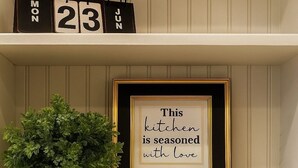 Uma prateleira com uma placa emoldurada que diz "Esta cozinha é temperada com amor", uma planta em vaso e maçãs e peras decorativas.