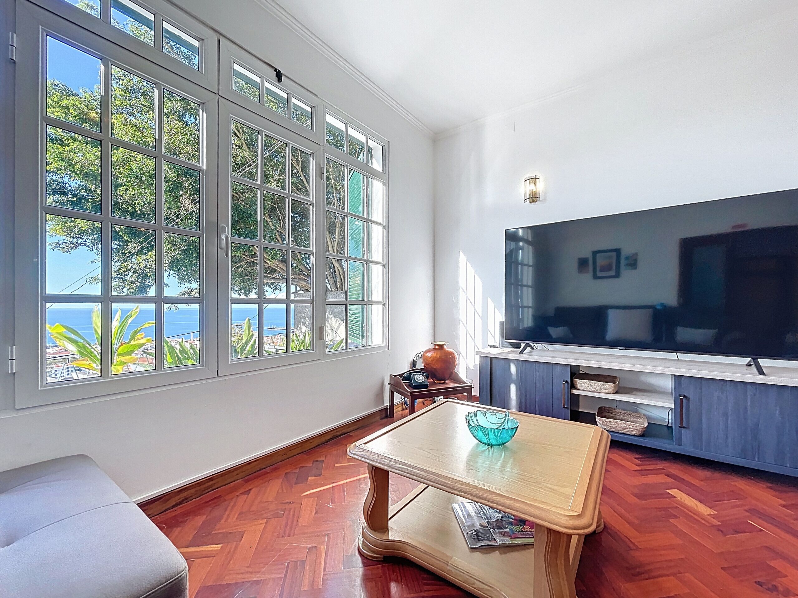 Un salón amplio con ventanas grandes, un televisor de pantalla plana, suelo de madera y una mesita con un cuenco.