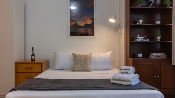 Una cama bien tendida con un plaid oscuro, una mesita de noche con lámpara, un aparador de madera con estantes, y un cuadro en la pared.