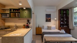 Una habitación de hotel compacta con kitchenette, una cama con banco, una mesita de noche pequeña, una mesita auxiliar, una estantería con adornos y una ventana con cortinas.