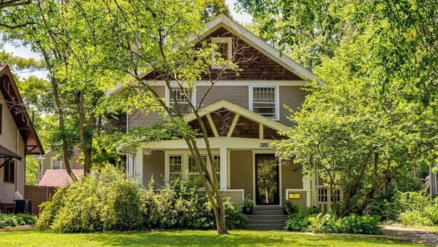 Kaksikerroksinen talo, jossa on etukuisti, ja sen ympärillä on vehreää kasvillisuutta ja puita.