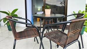 Pequeño patio al aire libre con sillas de mimbre y una mesa redonda, una planta en maceta y vistas del interior a través de una puerta de cristal.