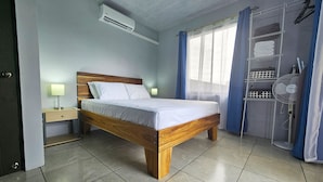 Un dormitorio con cama de madera, mesita de noche, aire acondicionado mural, ventana con cortinas de gasa y estante empotrado con toallas dobladas.