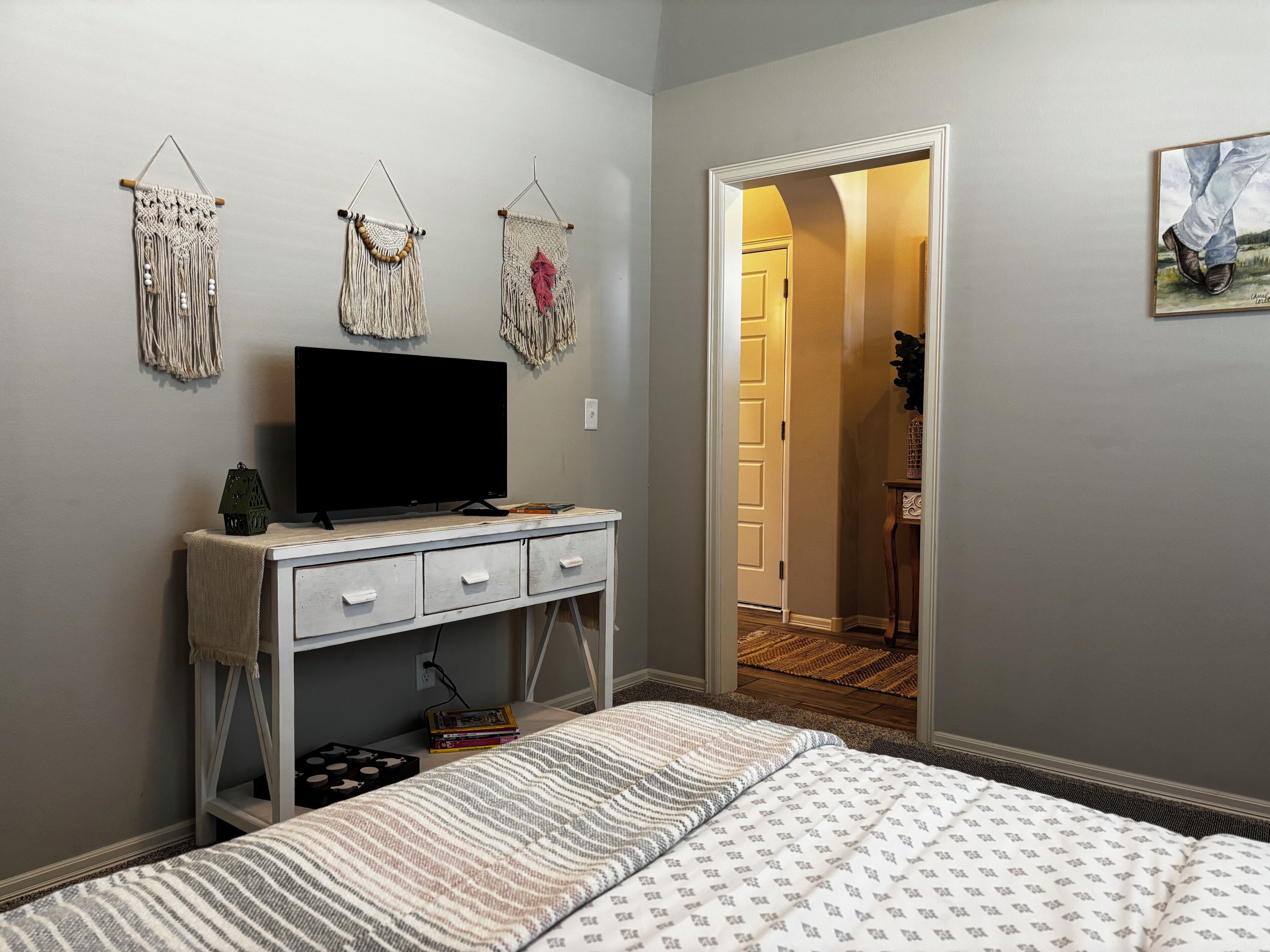 Een slaapkamer met een bed, een televisie op een dressoir en macramé wanddecoraties.