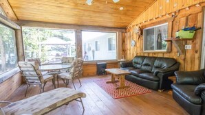 A cozy cabin interior with a wooden floor, a leather sofa, a dining table with chairs, and large windows offering a view of the outside. - Enjoy the Autumn colours on the edge of Algonquin park at Gramma's on the Lake (Barry's Bay)