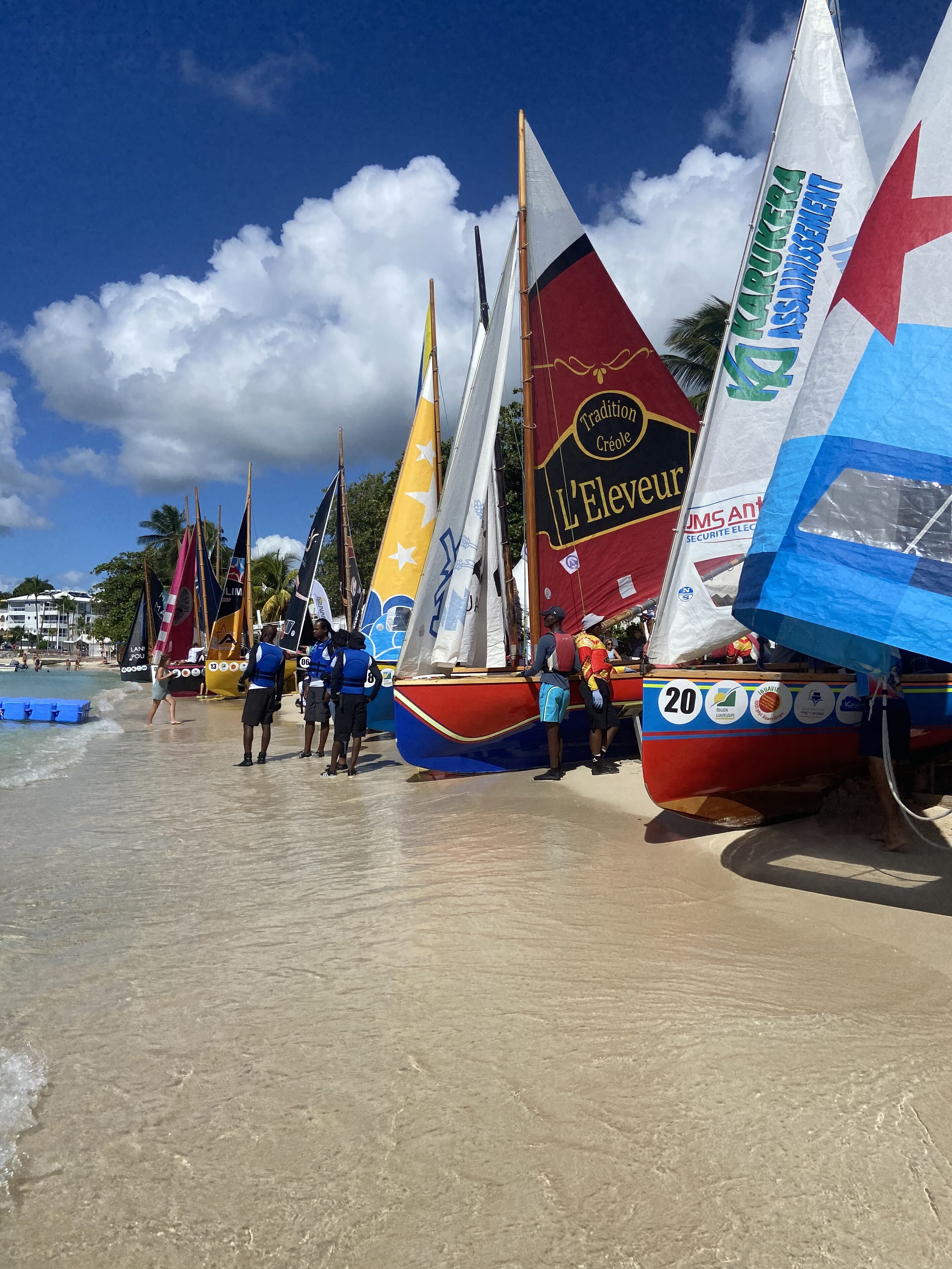 Une plage où des voiliers sont amarrés, des gens se tiennent debout aux alentours, et le ciel est dégagé.