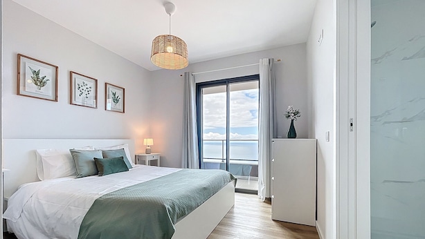 Une chambre à coucher avec un lit, une table de chevet, une lampe et une vue sur la mer, visible à travers une grande fenêtre.