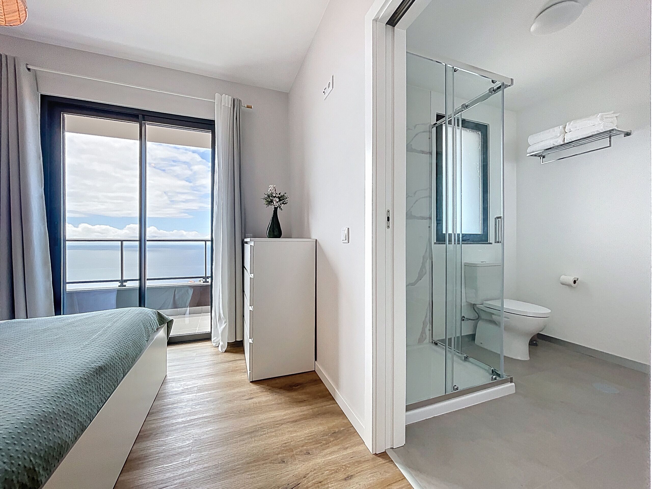 A hotel room with a bed, a small refrigerator, a glass-enclosed shower, a toilet, and a window with a view of the sea.