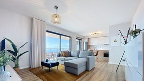 Un salon moderne avec un canapé gris, une table basse et une grande fenêtre donnant sur la mer.