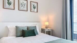 Un lit bien fait, avec une literie blanche et verte, une table de chevet avec une lampe, et trois reproductions botaniques encadrées accrochées au mur.
