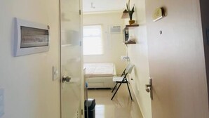A narrow hallway with a bed, a chair, a small table, and a trash bin.