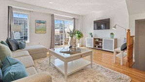 Ein modernes Wohnzimmer mit einer Couch, einem Couchtisch und einem Fernseher. Ein Fenster mit Vorhängen, eine Pflanze und ein Schrank mit Dekorationsobjekten sind ebenfalls zu sehen.