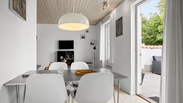 Un espace repas moderne avec une table, des chaises et un luminaire suspendu. Un téléviseur est fixé au mur et il y a une fenêtre avec des rideaux.