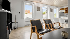 Un salon moderne comprenant un téléviseur, deux fauteuils en cuir noir, une table à manger et un coin cuisine avec des meubles en bois et un luminaire suspendu.