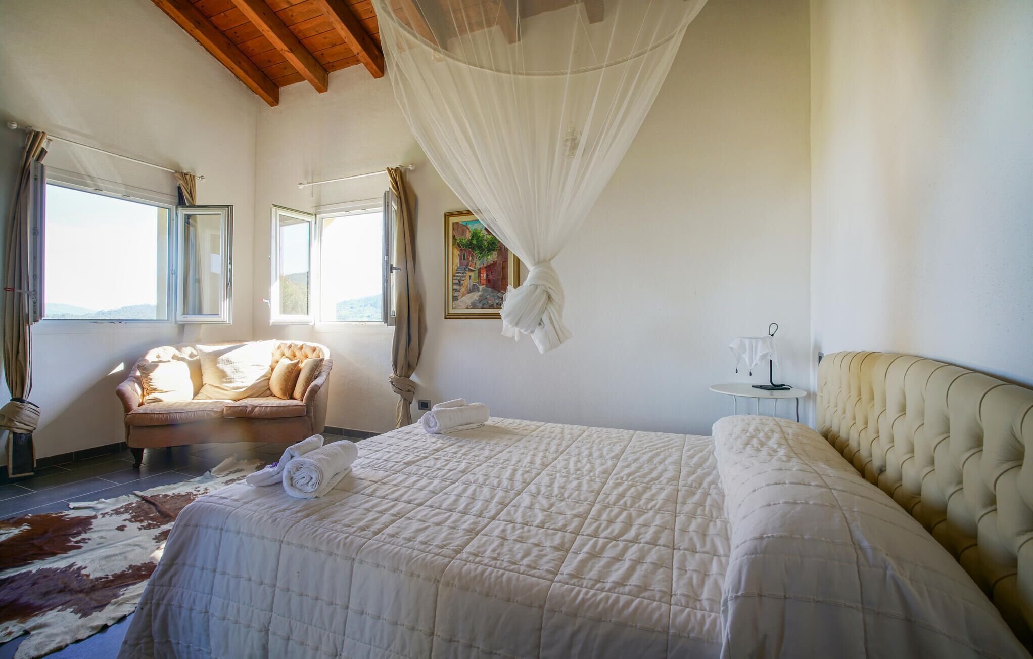 A bedroom with a large bed, a chaise lounge, a small table, and a painting on the wall.
