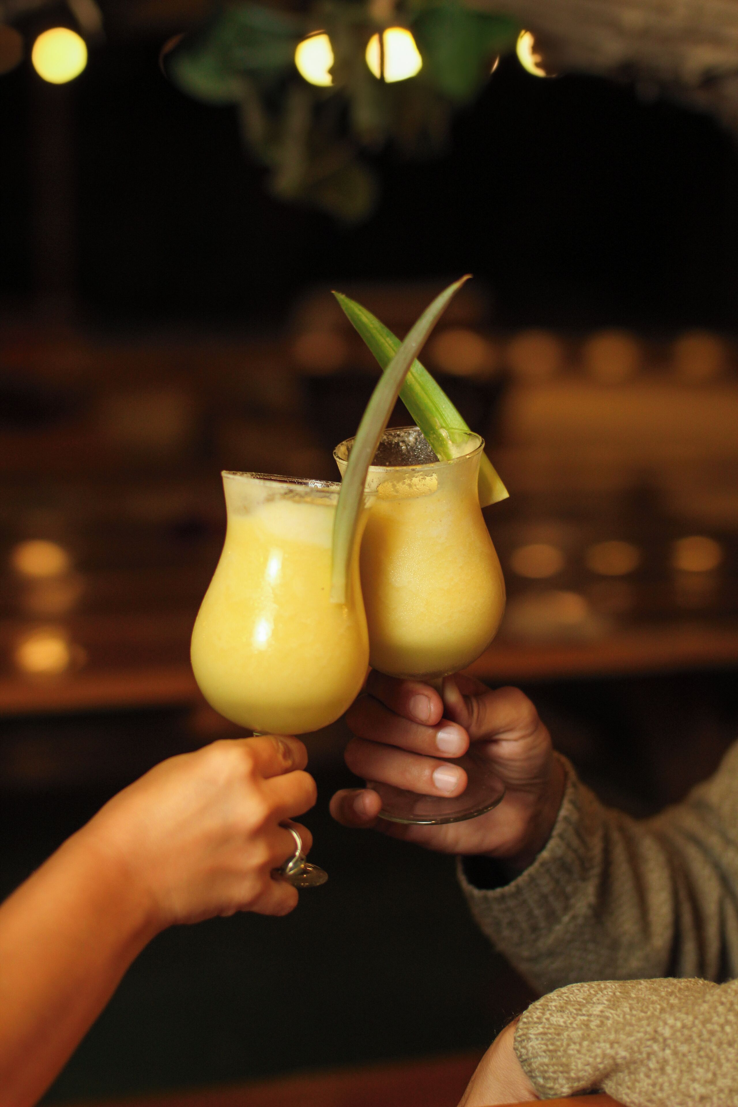 Two people holding glasses of a yellow beverage with straws, in a dimly lit indoor setting.