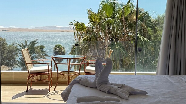 A bedroom with a large bed, a glass door leading to a balcony with a table and chairs, and a view of the sea.