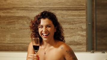 A person in a bathtub with a glass of red wine, a tattoo on the arm, and a necklace.