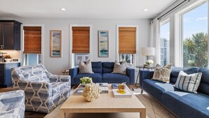 A modern living room with a blue sofa, patterned armchairs, a coffee table with books and a vase, and large windows with bamboo blinds.