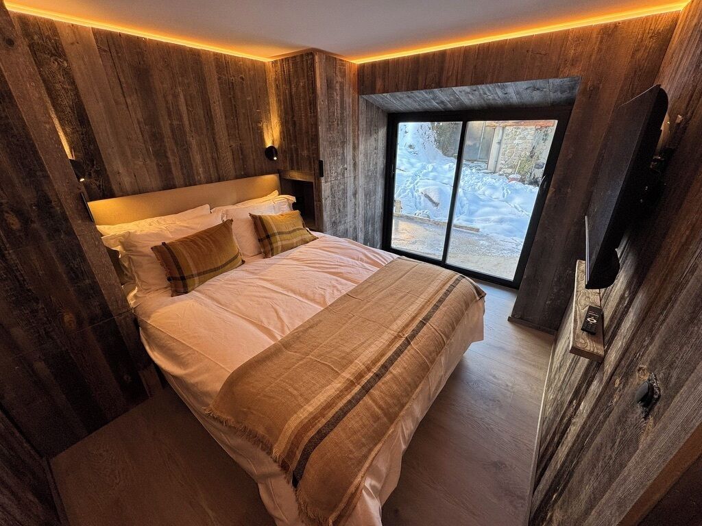 A bedroom with a large bed, wooden panel walls, a window with a view of snow, and a mounted TV.