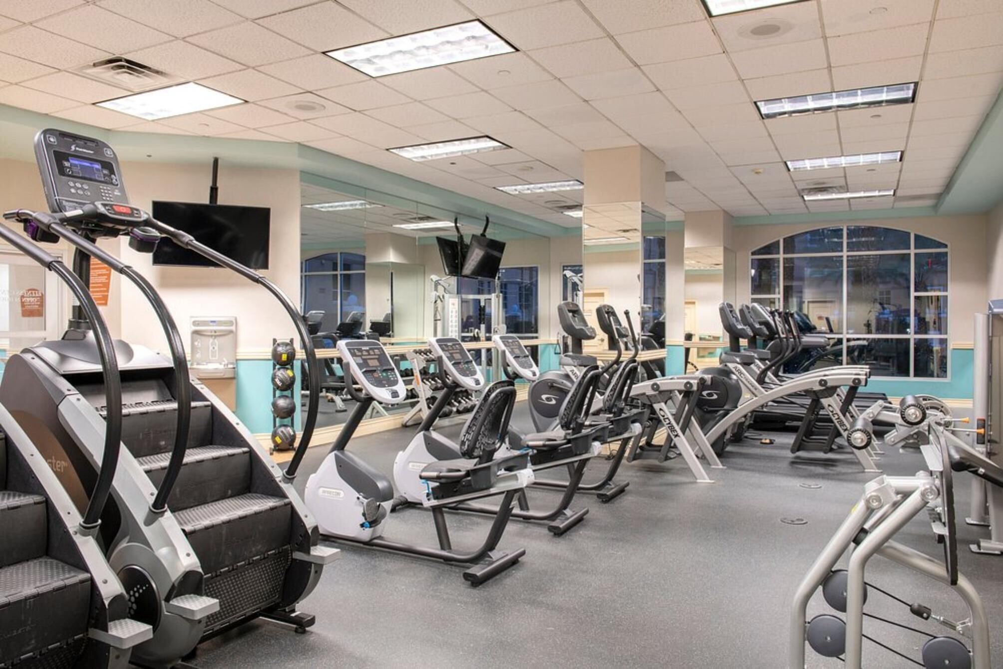 Un gimnasio con diferentes máquinas de cardio, espejos y un ventanal grande.
