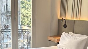 Et hotelværelse med en stor seng, et natbord, en væglampe og udsigt til en bygning og træer gennem et vindue.