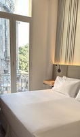 Habitación de hotel con una cama grande, una mesita de noche, una lámpara de pared y vistas a un edificio y árboles a través de una ventana.
