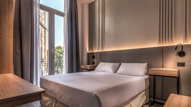 Et moderne hotelværelse med en stor seng, et trænatbord, et sengebord og udsigt til en bygning gennem et vindue.