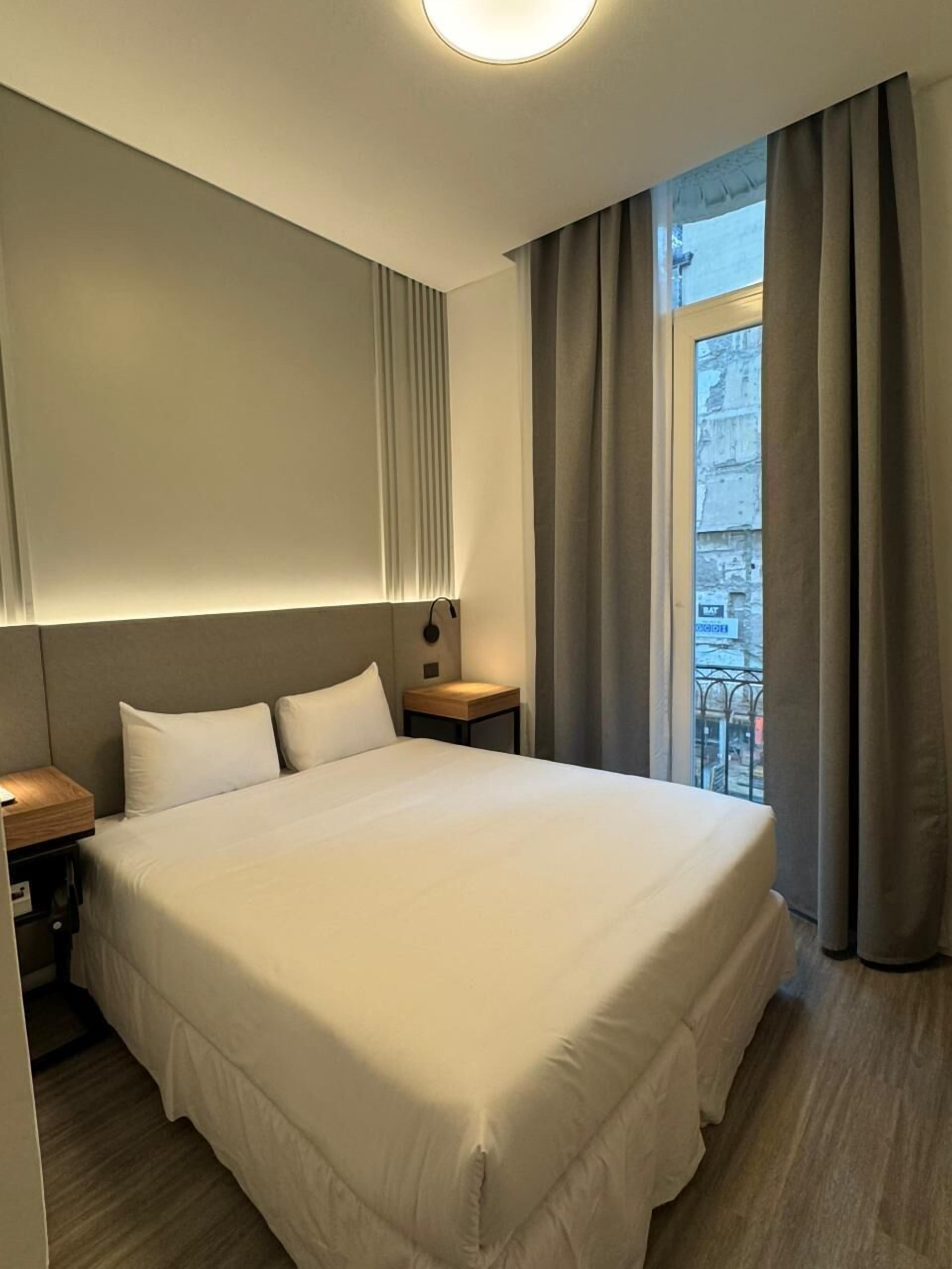 A modern hotel room with a large bed, a bedside table, a nightstand, and a window with curtains.