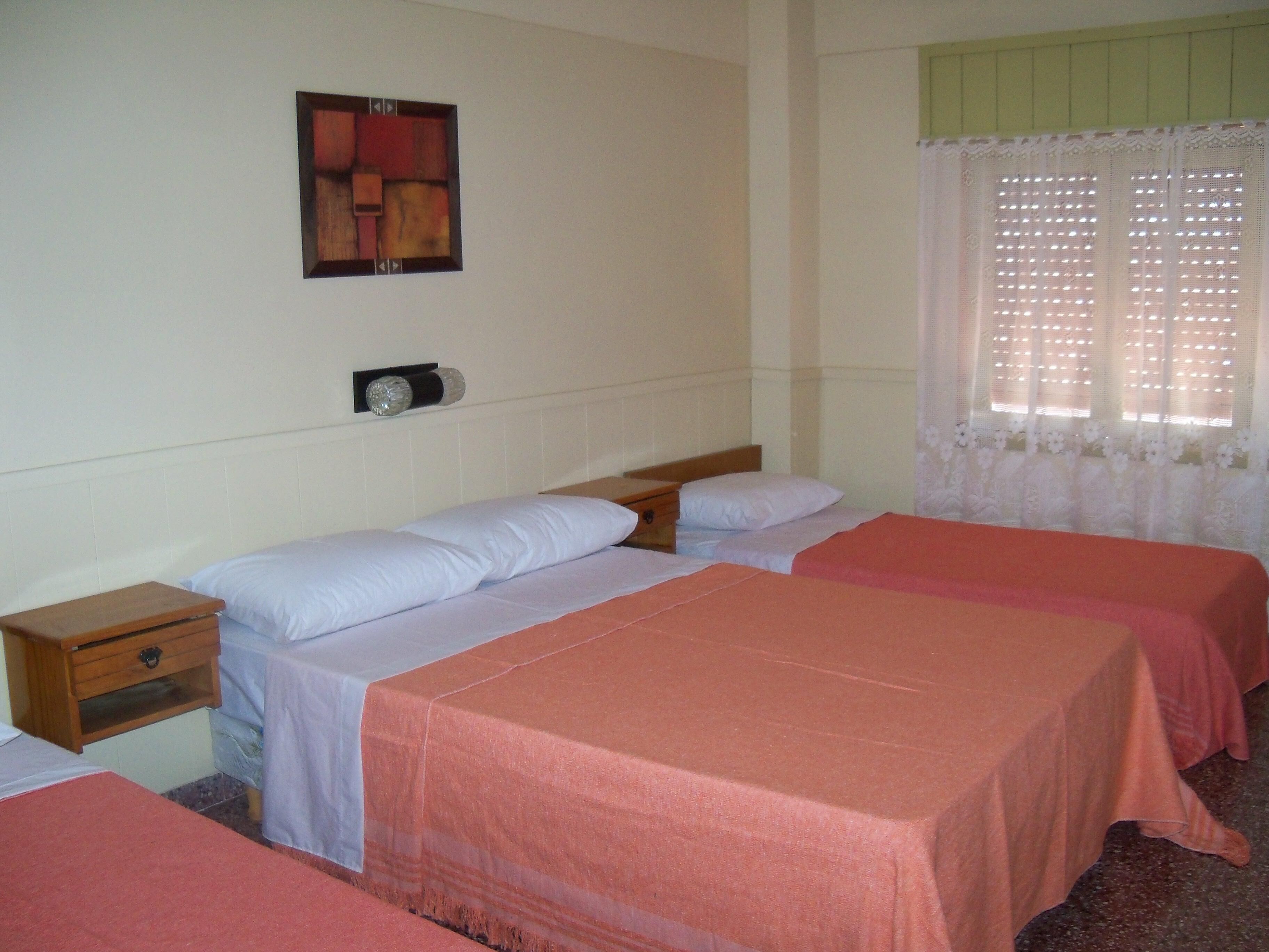 Une chambre d’hôtel avec deux lits simples, une table de chevet avec une lampe, une petite armoire en bois et une fenêtre avec des rideaux en dentelle.